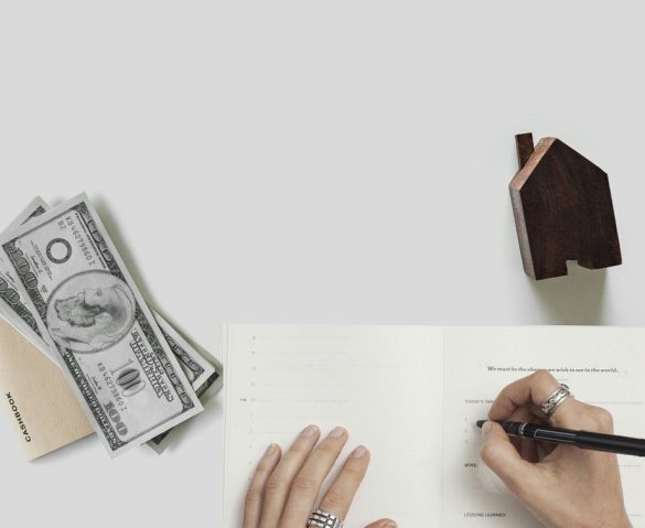 American 100 dollar bills with a notebook and wooden miniature house behind a grey background